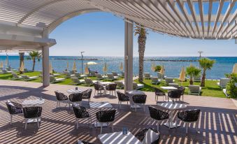 a beautiful outdoor terrace with umbrellas , tables , and lounge chairs , surrounded by a white ceiling and overlooking the ocean at Radisson Beach Resort Larnaca