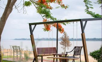 a wooden swing hanging from a tree , with a bench and table underneath it , in front of a body of water at Vieng Khong Hotel