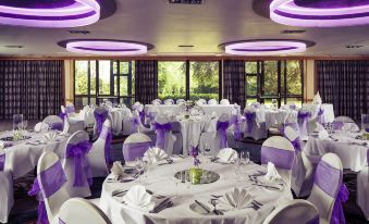 a large , well - lit banquet hall with tables covered in white tablecloths and chairs adorned with purple bows at Mercure Hull Grange Park Hotel