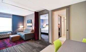 a modern hotel room with a couch , bed , and desk , as well as an open doorway leading to another room at Home2 Suites by Hilton Walpole Foxboro