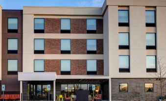 a modern hotel building with multiple floors , large windows , and outdoor seating areas under a clear blue sky at Home2 Suites by Hilton Dekalb