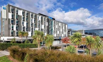 a modern hotel building with a large parking lot and palm trees in the foreground at Ibis Budget Auckland Airport