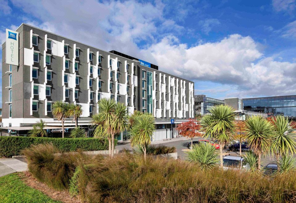 a modern hotel building with a large parking lot and palm trees in the foreground at Ibis Budget Auckland Airport