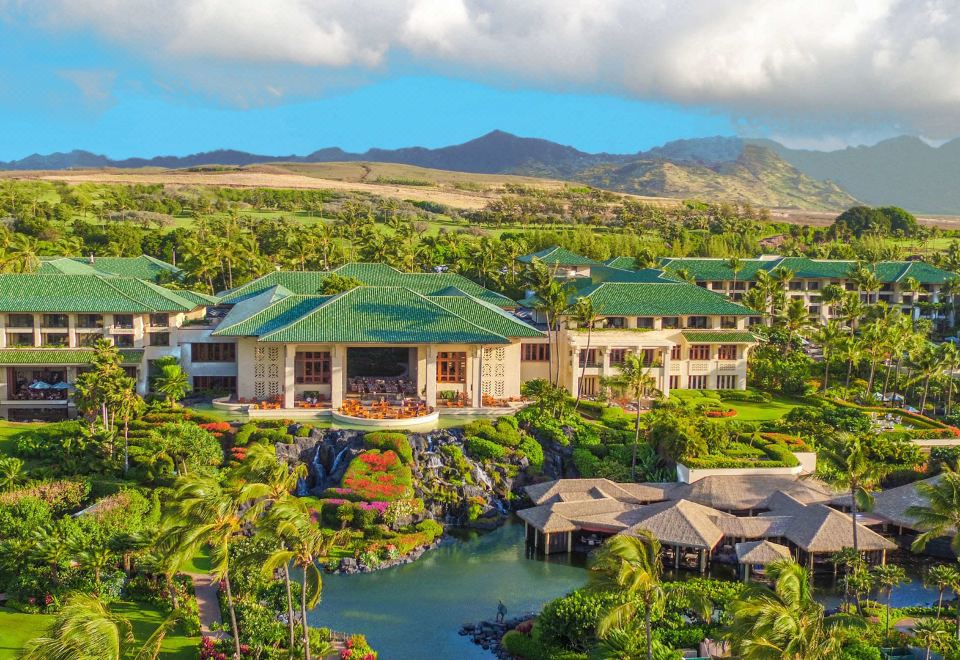 a large , multi - story building with a green roof is surrounded by lush greenery and mountains at Grand Hyatt Kauai Resort and Spa