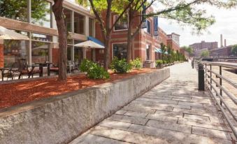 a brick sidewalk with trees and bushes lining the side , leading to a brick building at UMass Lowell Inn and Conference Center