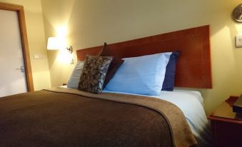 a bed with a brown blanket and two pillows , one blue and one brown , is illuminated by a wall - mounted lamp at The Corners Inn