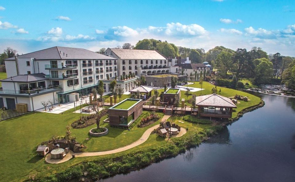 aerial view of a large hotel surrounded by lush green grass and trees , with a lake in the background at Galgorm