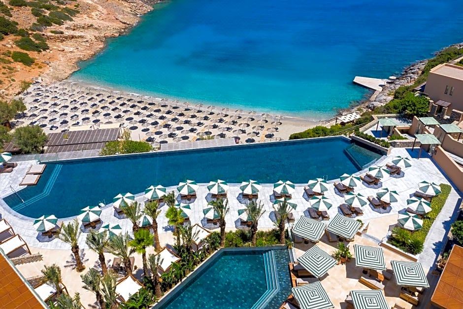 a large outdoor pool surrounded by lounge chairs and umbrellas , with a beach in the background at Daios Cove Luxury Resort & Villas