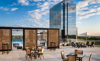 a rooftop terrace with wooden tables and chairs , overlooking a large skyscraper and a body of water at Renaissance Dallas at Plano Legacy West Hotel