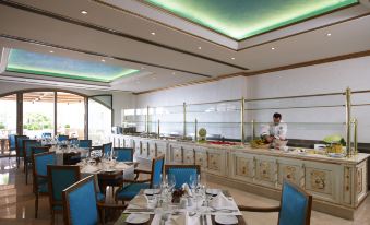 a large dining room with blue chairs and tables , a woman in a chef 's uniform working behind the counter at Mitsis Selection Laguna