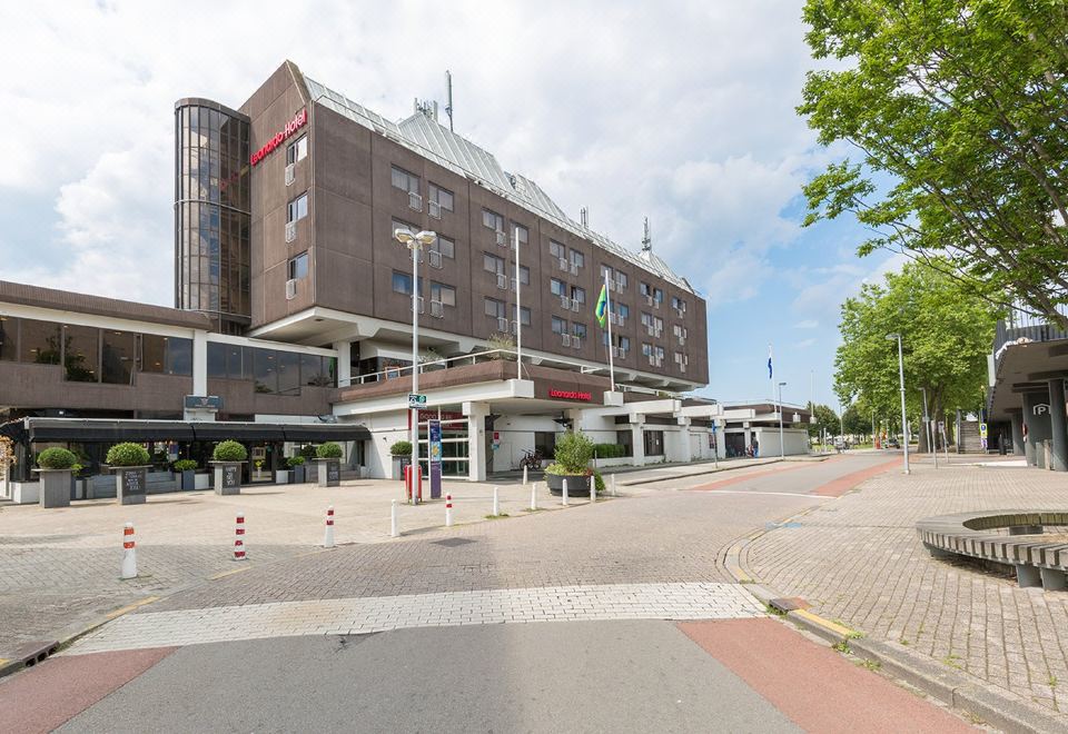 a large building with a red sign on the side is located next to a road at Leonardo Hotel Lelystad City Center
