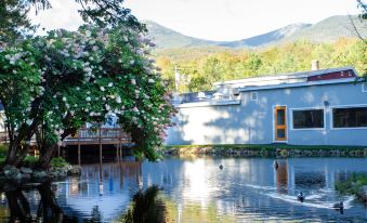 a house with a blue roof and white walls is surrounded by trees and flowers , reflected in the water of a lake at Woodwards White Mountain Resort, BW Signature Collection