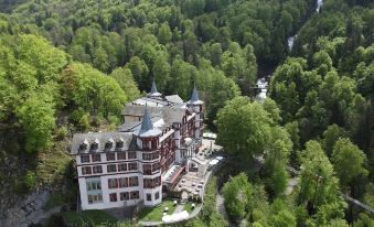 a large , ornate building with multiple turrets and a unique architectural design is surrounded by lush green trees at Grandhotel Giessbach
