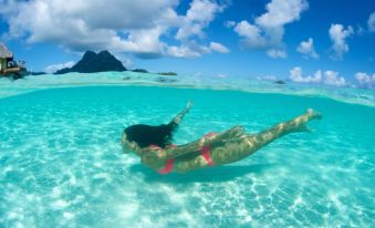 a woman is floating in the clear blue water near a mountain , enjoying the sunny day at Le Bora Bora by Pearl Resorts