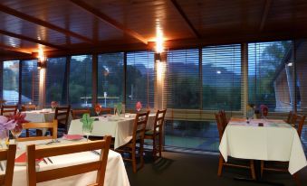 a restaurant with wooden tables and chairs , white tablecloths , and large windows offering a view of trees outside at Bucketts Way Motel