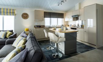 a modern kitchen and living room area with a gray couch , white bar stools , wooden ceiling , and wooden cabinets at King's Lynn Caravan & Camping Park