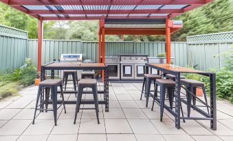 a backyard patio with a red pergola , outdoor dining tables , and stools arranged under the shade of trees at Black Forest Motel
