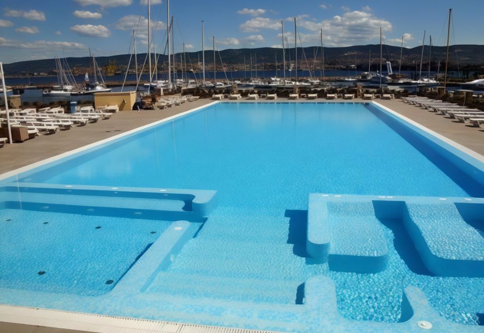 a large , empty swimming pool surrounded by lounge chairs and umbrellas , with boats in the background at Hotel San Rocco