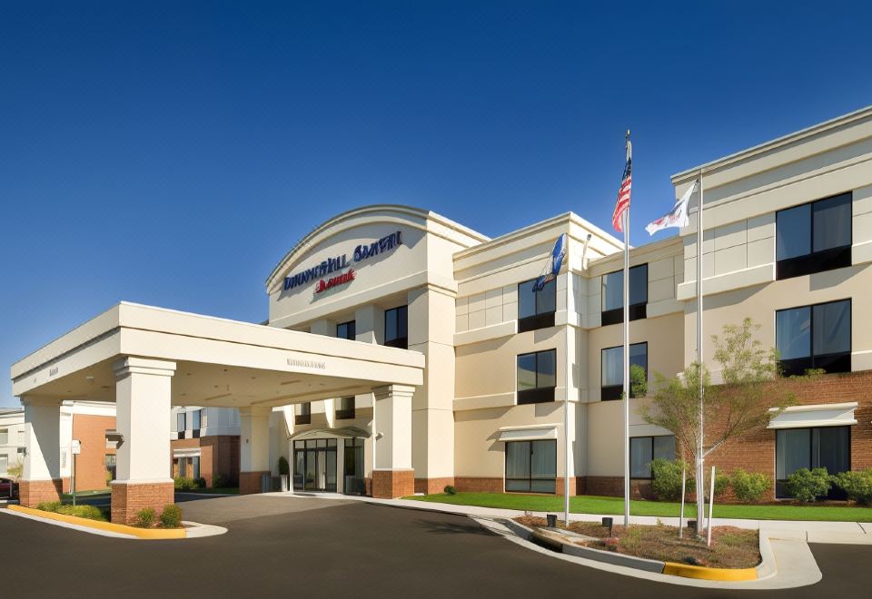 a large , white hotel building with a covered entrance and american flags flying outside , under a clear blue sky at SpringHill Suites Alexandria