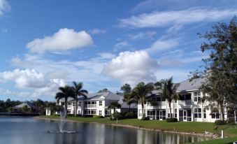 a serene scene of a lake surrounded by white buildings with palm trees and clouds in the sky at GreenLinks Golf Villas at Lely Resort