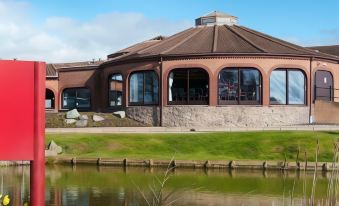 a large , modern building with a brick exterior and large windows is surrounded by water and greenery at Leonardo Hotel and Conference Venue Hinckley Island