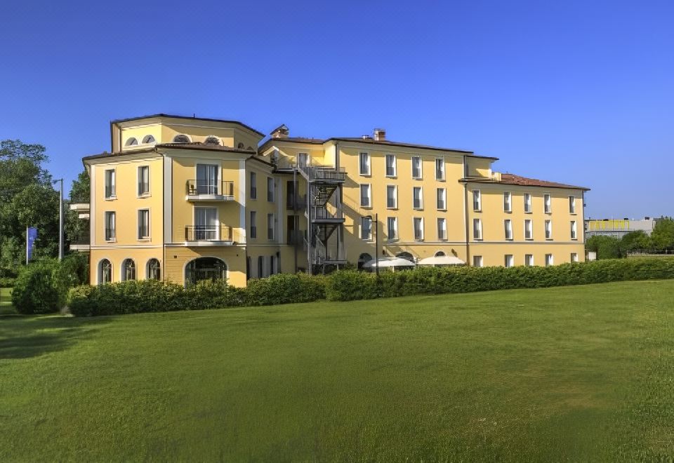 a large yellow building surrounded by green grass and trees , with a blue sky in the background at Maranello Palace Hotel