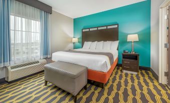 a large bed with white linens and a brown headboard is in the center of a room with yellow and gray striped carpet at La Quinta Inn & Suites by Wyndham Houston Humble Atascocita