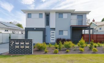 a two - story house with a blue and white exterior is surrounded by greenery , including bushes and flowers at East Maitland Executive Apartments