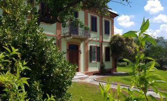 a two - story house with green shutters and a red roof , surrounded by trees and bushes , in a sunny day at La Fornace