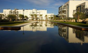 a large , well - maintained pool of water is surrounded by white buildings and palm trees , with the sun shining through clouds at El Plantío Golf Resort