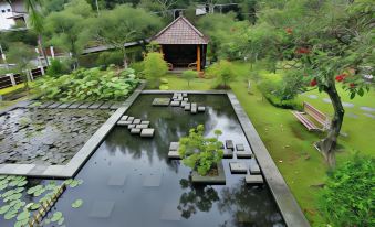 a serene garden with a pond filled with floating objects , surrounded by lush greenery and a small wooden hut at Hotel Cianjur Cipanas