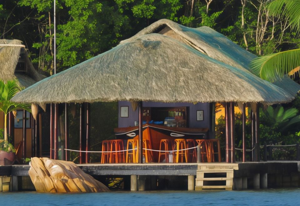 a thatched - roof hut with a dining table and chairs on a wooden platform , surrounded by water and trees at Whale Island Resort