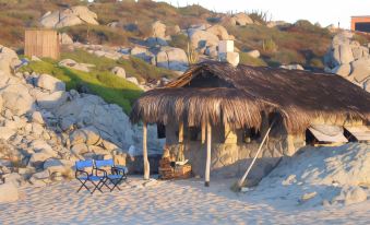 a small hut with a thatched roof is nestled on a sandy beach , surrounded by rocks and plants at Villa del Faro