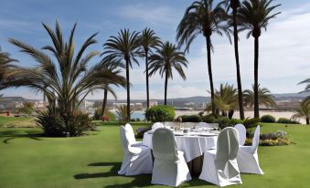 a dining table set up on a grassy lawn , surrounded by palm trees and a beautiful view of the ocean at Parador de Javea