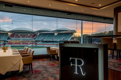 "a restaurant with a large window overlooking a sports stadium and chairs , with the logo of "" r "" in the background" at Oval Hotel at Adelaide Oval, an EVT hotel