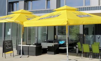 a patio area with yellow umbrellas and couches , providing shade for people and equipment , under a sunny sky at Beachin - Sport, Events, Hotel, Restaurant, Bar