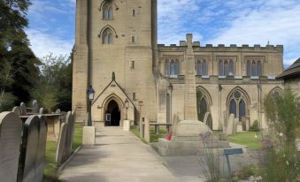 a large stone church with a clock tower , surrounded by green grass and trees , under a blue sky with clouds at The Green Dragon at Bedale