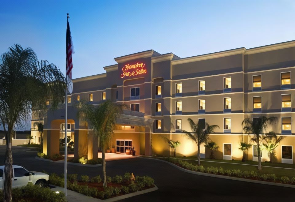 a hampton inn hotel with its sign lit up , surrounded by palm trees and a flag in the foreground at Hampton Inn & Suites Lake Wales