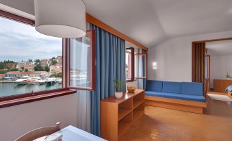 a cozy living room with wooden furniture , blue curtains , and a view of the water through large windows at Maistra Select Riva Apartments