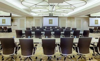 a large conference room with multiple rows of chairs arranged in front of a projector screen , ready for a meeting at The H Hotel