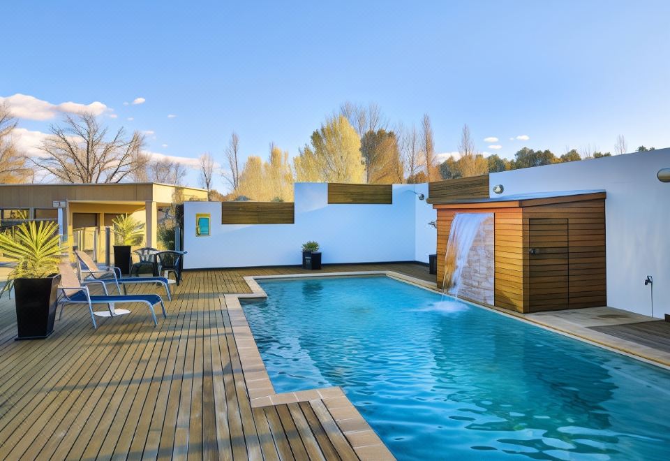 a modern swimming pool area with a large wooden deck and water feature , surrounded by trees and buildings at Myrtleford Motel on Alpine