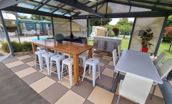 a wooden dining table surrounded by white stools and chairs in an outdoor setting , with a pool visible in the background at Lone Pine Motel