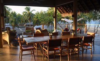 a large wooden dining table surrounded by chairs on a covered patio , with a view of the ocean in the background at Karpaha Sands
