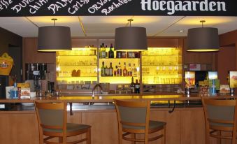 a bar with wooden chairs and tables , a neon sign above the counter , and various bottles and glasses on display at Ibis Brussels Airport