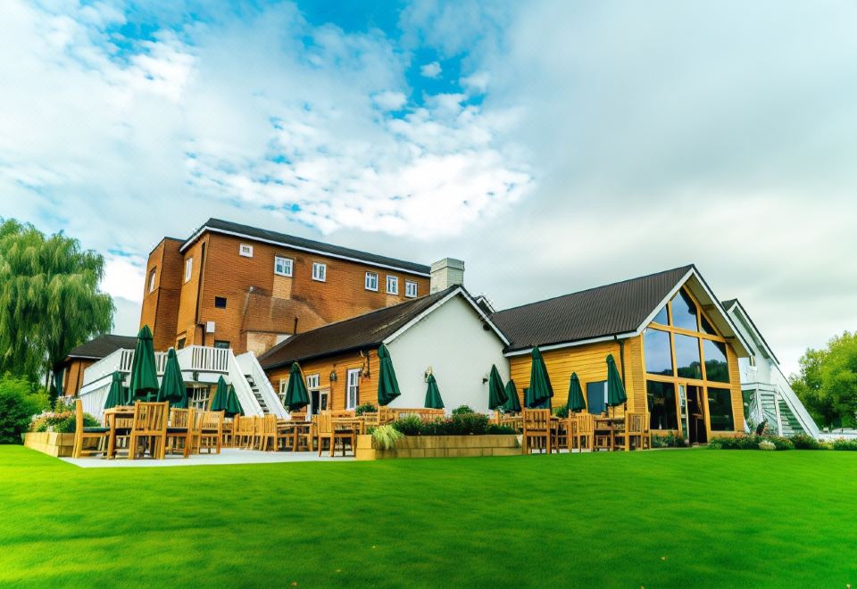 a large , yellow building with green umbrellas and tables set up in front of it , set against a cloudy sky at Arrow Mill- Brunning and Price