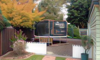 a small bus is parked in a grassy area next to a white picket fence , surrounded by trees and bushes at Asure Green Gables Motel