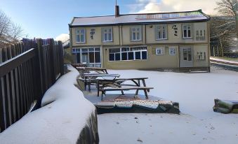 a snow - covered yard with a large building and several picnic tables , surrounded by a wooden fence at Anglers Arms