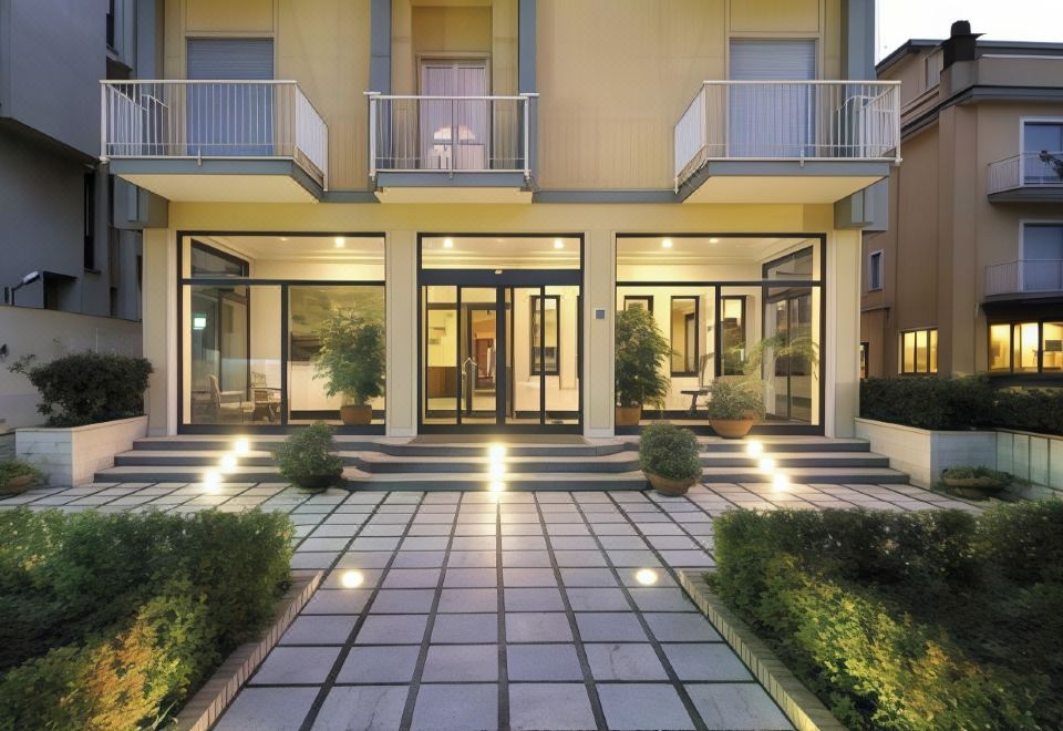 a modern , two - story building with multiple floors and large windows , surrounded by greenery and lit up by lights on the ground at American Hotel