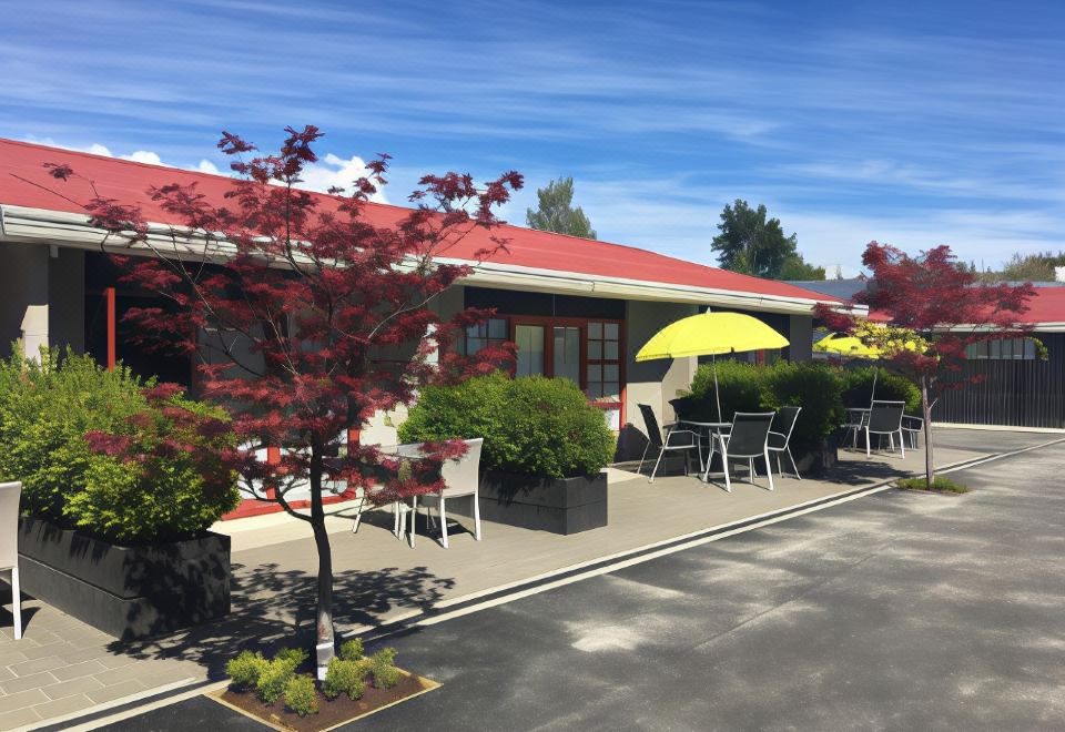 a red - roofed building with a yellow umbrella and tables and chairs outside , surrounded by trees and a parking lot at Arrowtown Motel