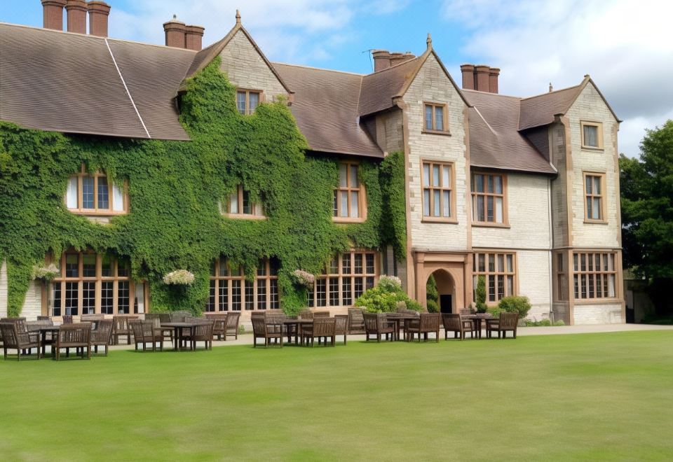 a large house with a green ivy - covered roof is surrounded by a lawn and has several chairs placed on the lawn at The Billesley Manor Hotel